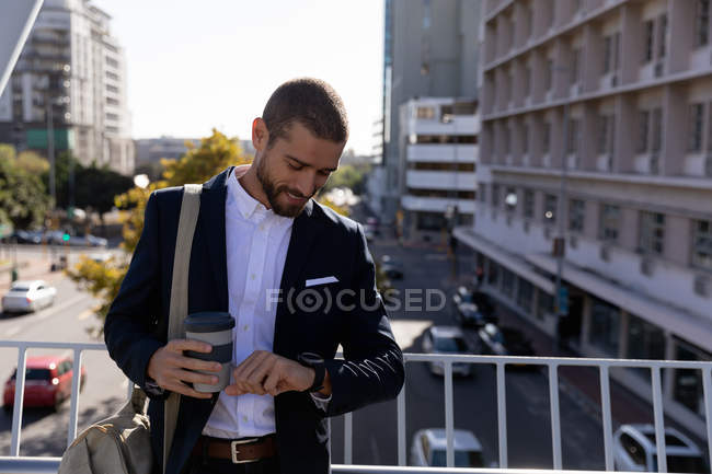 Вид спереди улыбающегося молодого кавказца, держащего кофе на вынос и проверяющего время на часах, стоящего на дорожке по городской дороге. Цифровая реклама на ходу . — стоковое фото