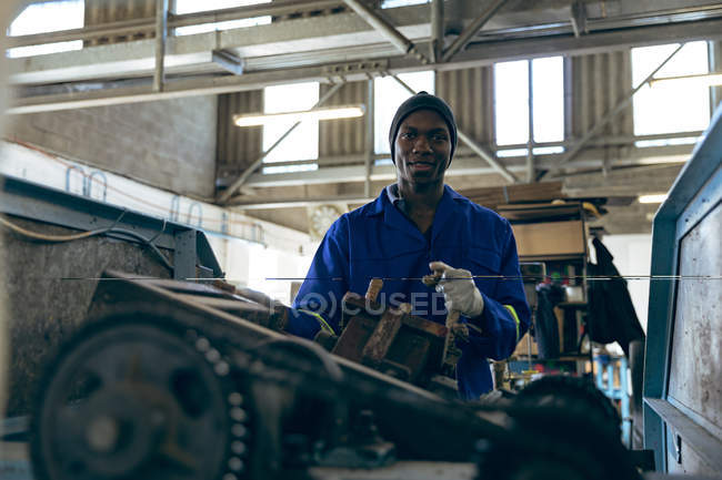 Portrait of a young African American man wearing overalls and a hat operating a machine at a cricket ball factory and smiling to camera. — Stock Photo
