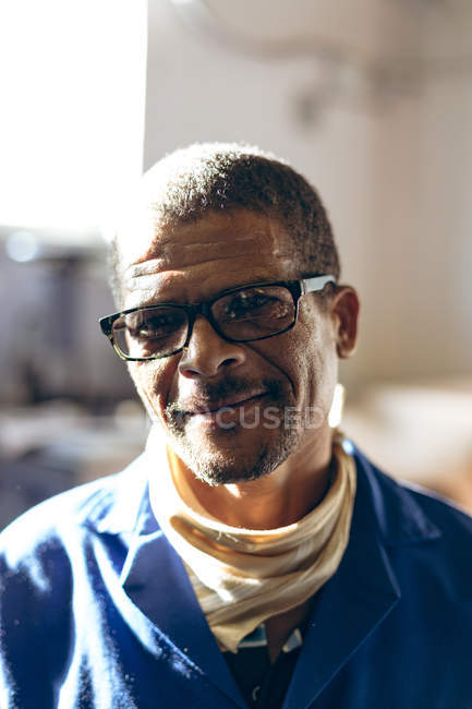 Portrait close up of a middle aged mixed race man wearing glasses, looking to camera and smiling at a factory making cricket balls where he works. — Stock Photo