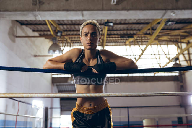 Low angle view of female boxer leaning on ropes and looking at camera in boxing ring. Strong female fighter in boxing gym training hard. — Stock Photo