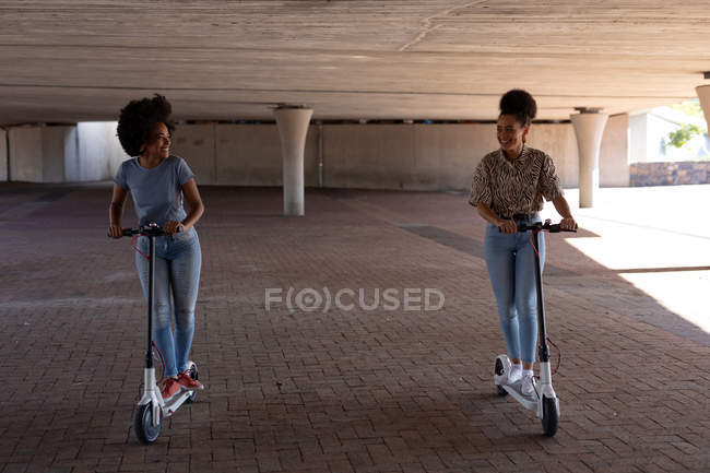 Vista frontal de dos hermanas adultas jóvenes de raza mixta montadas en scooters eléctricos en un parque urbano, mirándose sonrientes - foto de stock