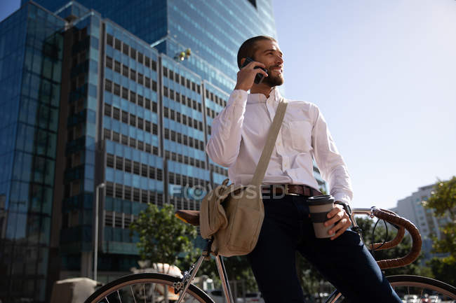 Vista frontal de perto de um jovem caucasiano segurando um café takeaway e falando em um smartphone, apoiando-se em sua bicicleta em uma rua da cidade. Digital Nomad em movimento . — Fotografia de Stock