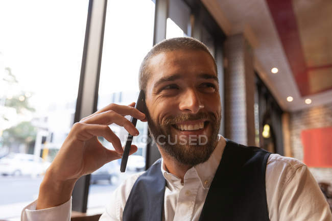 Vista frontal de cerca de un joven caucásico sonriente en una llamada telefónica sentado en una mesa dentro de un café, mirando hacia otro lado. Nómada digital en movimiento . - foto de stock