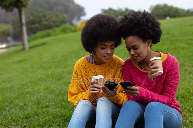 Vista frontal de cerca de dos hermanas adultas jóvenes de raza mixta sonriendo, mirando sus teléfonos inteligentes y sosteniendo cafés para llevar, sentadas juntas en la hierba en un parque - foto de stock