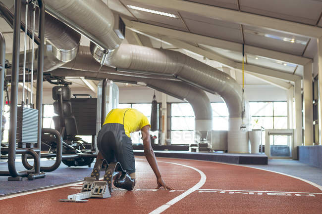 Vista trasera del atlético masculino afroamericano discapacitado en el bloque de partida en la pista de atletismo en el gimnasio - foto de stock