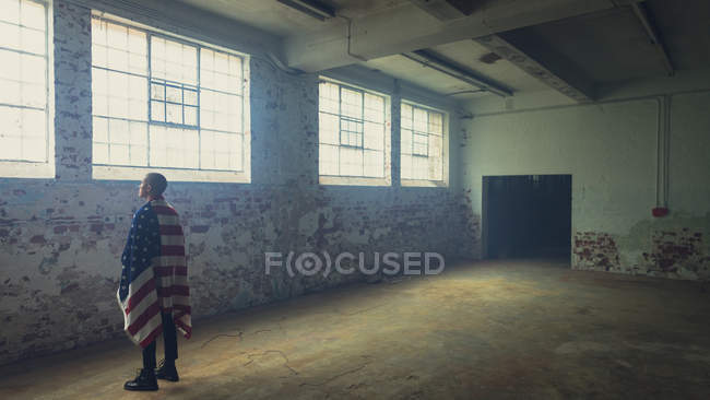 Seitenansicht eines jungen hispanisch-amerikanischen Mannes mit einer amerikanischen Flagge über den Schultern, der in einer leeren Lagerhalle steht und zum Fenster blickt — Stockfoto