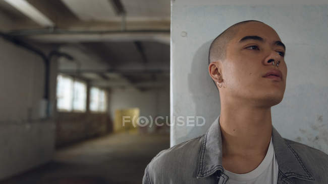 Front view of a young Hispanic-American man with piercings wearing a plain white shirt inside a grey jacket against a white wall inside an empty warehouse looking away from the camera — Stock Photo
