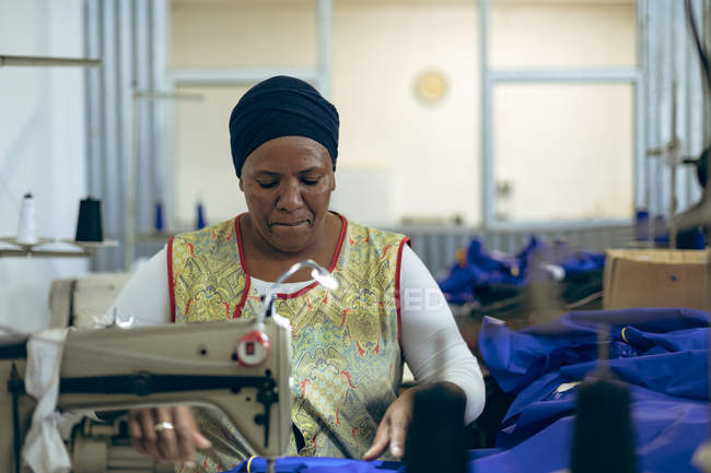Вид спереду крупним планом жінка середнього віку змішаної раси, яка сидить і керує швейною машиною на заводі спортивного одягу . — стокове фото