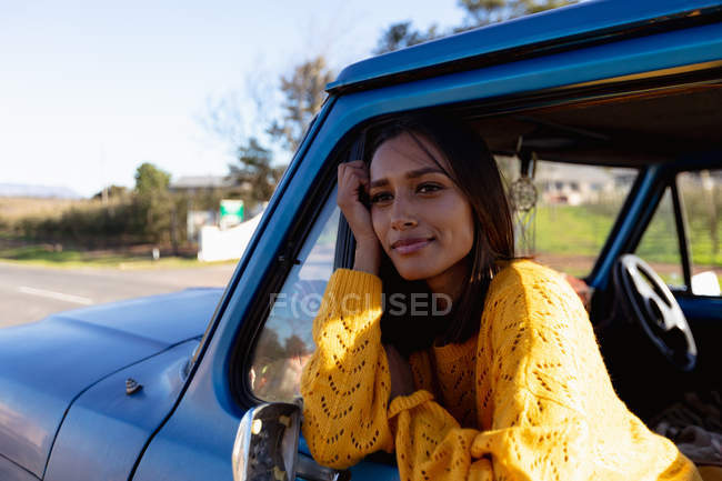Portrait d'une jeune femme métisse assise sur le siège passager avant d'une camionnette, penchée par la fenêtre latérale souriante lors d'un voyage en voiture — Photo de stock