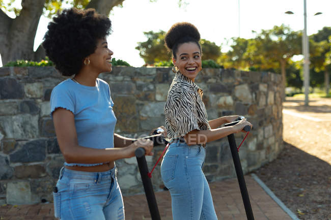Вид сбоку на двух взрослых сестер смешанной расы, катающихся на электрических скутерах в городском парке, улыбающихся друг другу — стоковое фото
