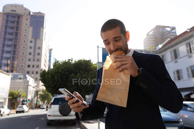 Вид спереди на молодого кавказца, использующего смартфон и поедающего выпечку в бумажном пакете, стоящего на городской улице. Цифровая реклама на ходу . — стоковое фото