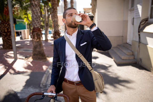 Vista frontal de un joven caucásico tomando un café para llevar, de pie con su bicicleta en una calle de la ciudad. Nómada digital en movimiento . - foto de stock