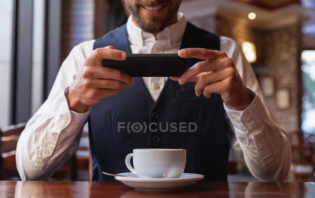 Vue de face au milieu d'un jeune homme caucasien souriant utilisant son smartphone assis à une table avec une tasse de café à l'intérieur d'un café. Nomade numérique en mouvement . — Photo de stock