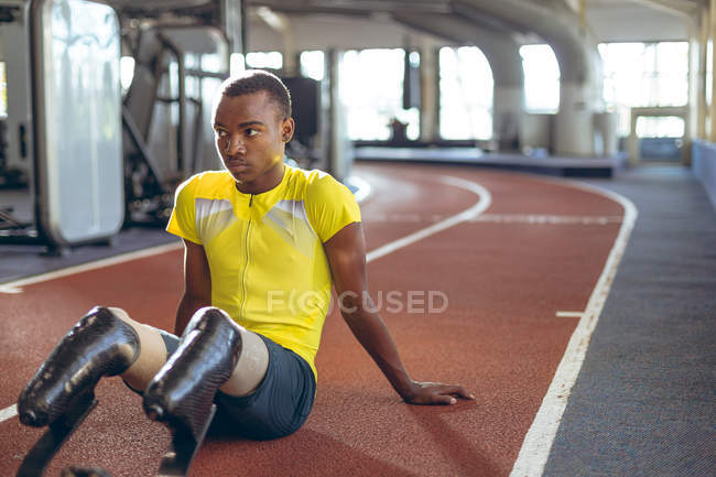 Vista frontal de un atlético afroamericano discapacitado relajándose en una pista de atletismo en el gimnasio - foto de stock