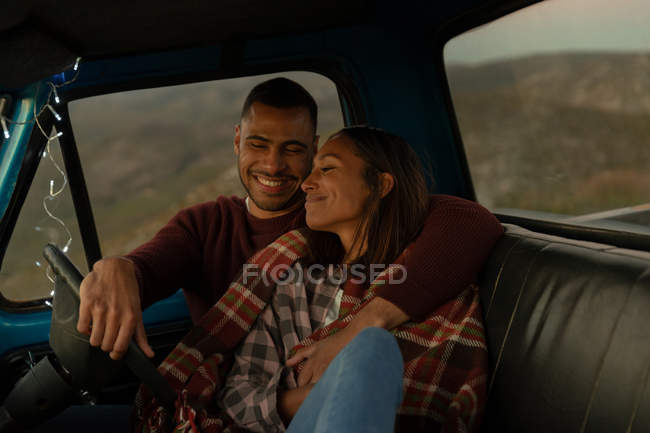 Vista frontal de una joven pareja mixta sentada en su camioneta, sonriendo y abrazándose al atardecer durante una parada en un viaje por carretera. Están sentados en los asientos delanteros y el interior del coche está iluminado con luces de cuerda . - foto de stock