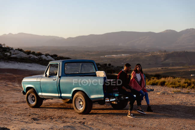 Vista lateral de um jovem casal misto sentado na parte de trás de seu caminhão pick-up conversando durante uma parada em uma viagem de carro . — Fotografia de Stock