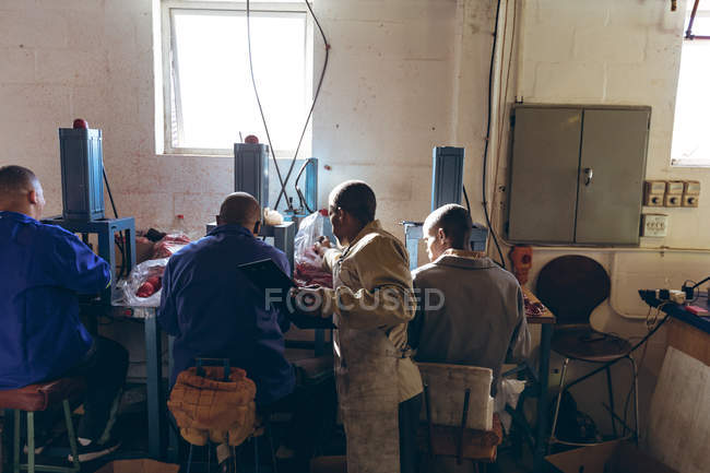 Rückansicht eines jungen afrikanisch-amerikanischen Mannes mit Klemmbrett, der hinter einer bunt gemischten Gruppe männlicher Arbeiter steht, die Maschinen bedienen und in der Werkstatt einer Cricketballfabrik das Fließband kontrollieren.. — Stockfoto