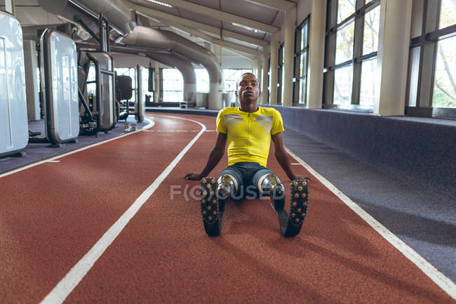 Vista frontal de un atlético afroamericano discapacitado relajándose en una pista de atletismo en el gimnasio - foto de stock