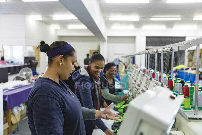 Side view of two young mixed race women talking a they check and operate automated sewing machines stitching shirts in a sports clothing factory, in the background are other colleagues also working with the machines. — Stock Photo