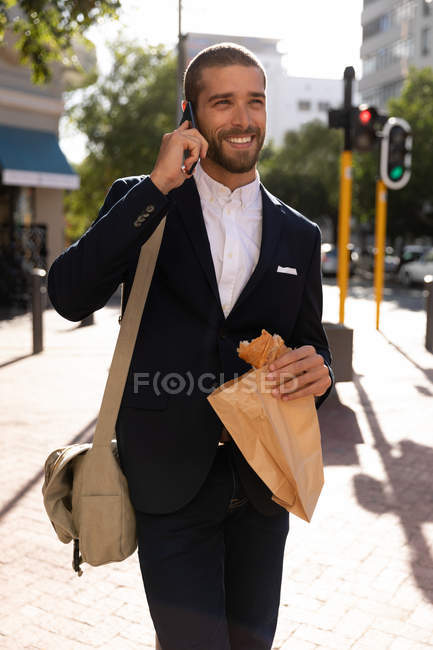 Vista frontale di un giovane caucasico sorridente che parla sullo smartphone e tiene una pasticceria in un sacchetto di carta mentre cammina in una strada della città. Nomade digitale in movimento . — Foto stock