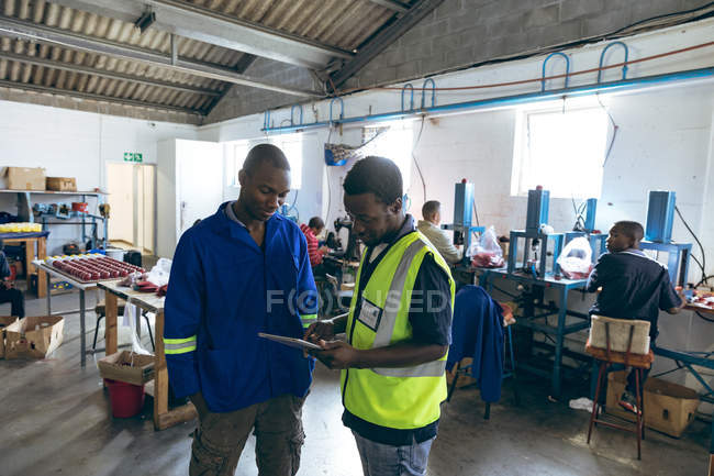 Front view of two young African American male colleagues at a cricket ball factory standing, talking and looking at a tablet computer, while people work operating machines in the background. — Stock Photo