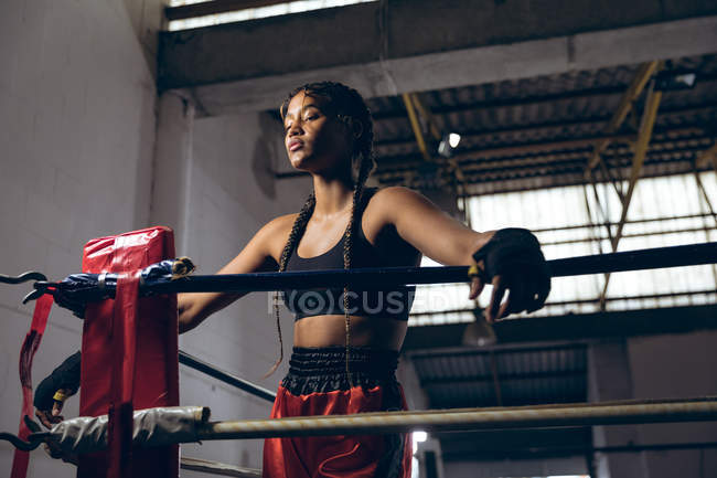 Низкоугольный вид женщины-боксера с закрытыми глазами, опирающейся на веревки в боксерском ринге в боксерском клубе. Сильная женщина-боец в боксёрском зале тяжело тренируется . — стоковое фото