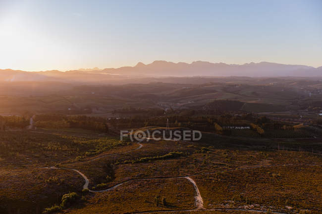 Vista ampla de uma paisagem natural aberta com trilhas para montanhas distantes e pôr do sol no horizonte — Fotografia de Stock