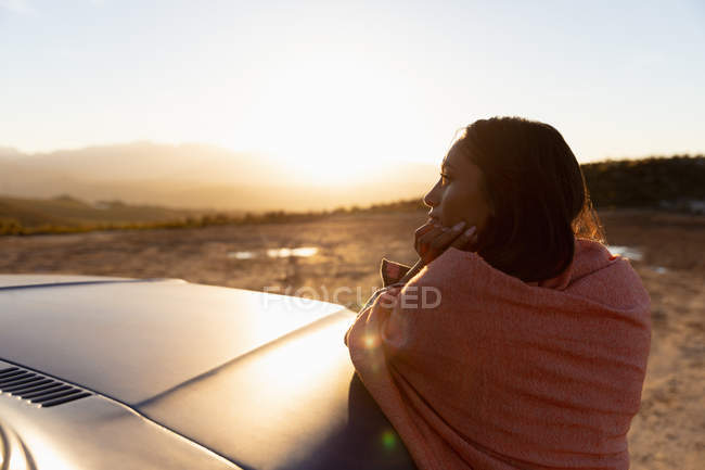 Крупним планом вид на молоду змішану жінку, що спирається на капот пікапа і насолоджується видом на світанку під час зупинки на сільській дорозі — стокове фото