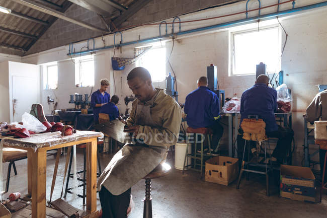 Seitenansicht eines jungen afrikanisch-amerikanischen Mannes, der auf einem Hocker sitzt und auf einem Klemmbrett in der Werkstatt einer Cricketballfabrik schreibt. — Stockfoto