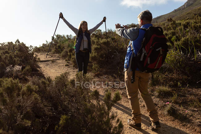 Rear view of a mature Caucasian man taking a photo of his partner, a mature Caucasian woman, standing on a trail and raising her Nordic walking sticks in the air during a hike — Stock Photo