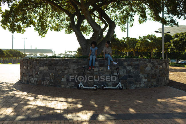 Вид спереди на двух молодых улыбающихся взрослых сестер смешанной расы, сидящих на стене в городском парке, с электрическими скутерами, припаркованными под ними — стоковое фото