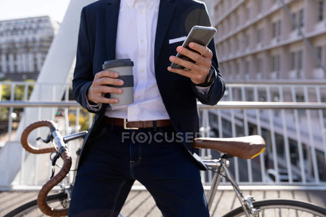 Вид спереду середина людини, що тримає винос кави і за допомогою смартфона, спираючись на велосипеді на доріжці над міською дорогою. Цифрові кочівники на ходу. — стокове фото