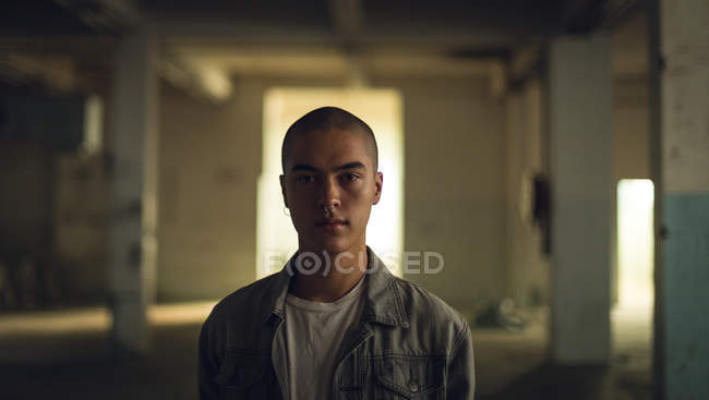 Vista frontale di un giovane ispano-americano con piercing che indossa una giacca grigia sopra una camicia bianca guardando attentamente la telecamera all'interno di un magazzino vuoto — Foto stock
