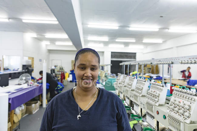 Porträt einer jungen Mischlingshündin, die in einer hell erleuchteten Sportbekleidungsfabrik neben Maschinenreihen steht, in die Kamera blickt und lächelt. Im Hintergrund sind einige Mitarbeiter zu sehen. — Stockfoto
