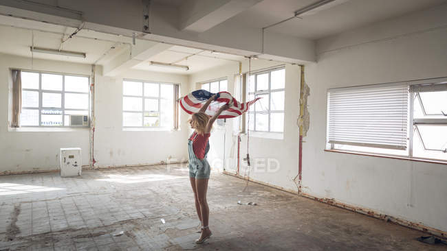 Vista lateral de una joven mujer caucásica sosteniendo una bandera estadounidense dentro de un almacén vacío - foto de stock