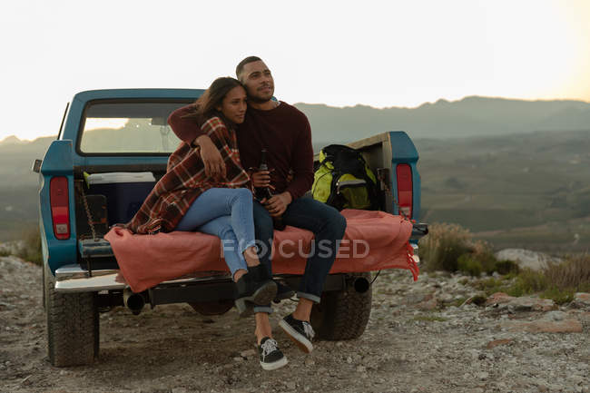 Vista frontal de una joven pareja mixta sentada en la parte trasera de su camioneta, abrazando y disfrutando de la vista al atardecer durante una parada en un viaje por carretera . - foto de stock