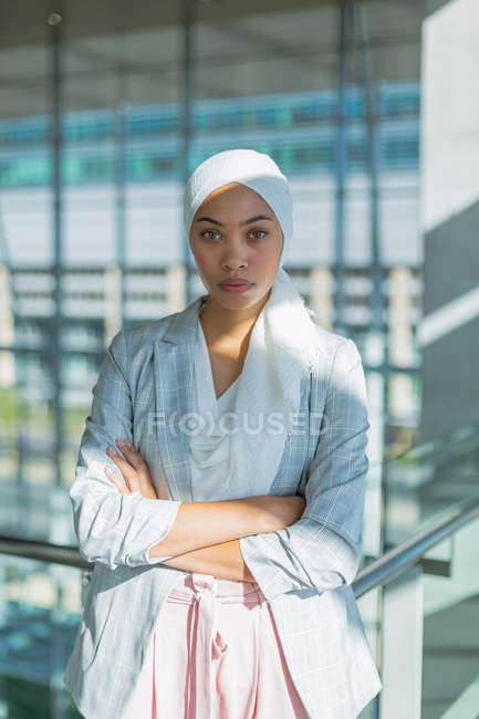 Close-up de empresária no hijab com os braços cruzados olhando para a câmera no corredor no escritório moderno . — Fotografia de Stock