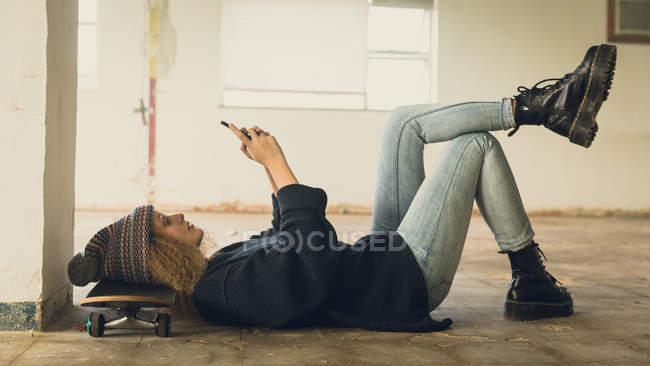 Vue latérale d'une jeune femme caucasienne aux cheveux bouclés portant une chemise noire et un bonnet couché sur le sol avec la tête sur un skateboard tout en utilisant un téléphone portable — Photo de stock