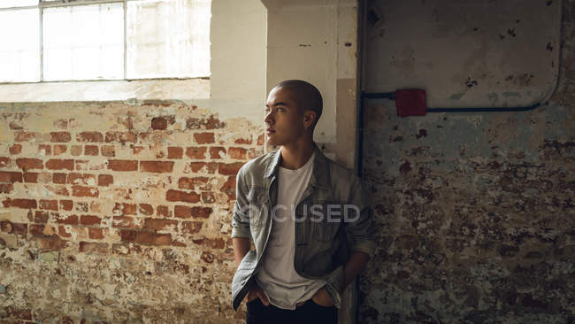 Frontansicht eines jungen hispanisch-amerikanischen Mannes, der eine graue Jacke über einem weißen Hemd trägt und mit den Händen in den Taschen von der Kamera wegschaut, während er in einer leeren Lagerhalle an der Wand steht — Stockfoto
