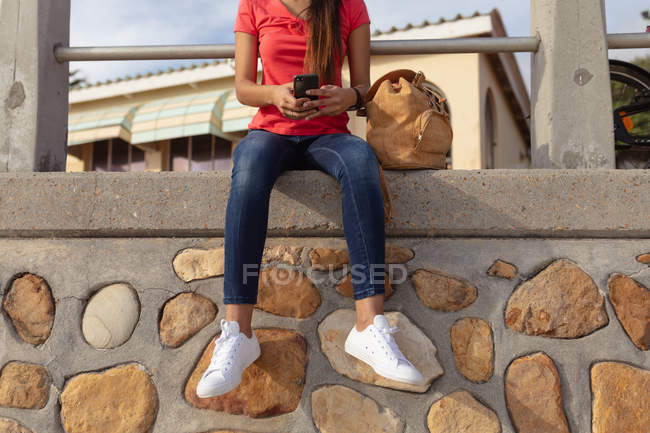 Вид спереду низька частина жінки, що сидить на вулиці на стіні з сумкою на сонці за допомогою смартфона — стокове фото