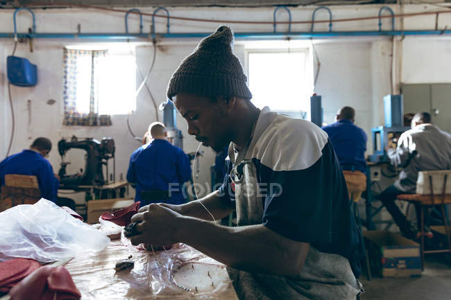 Side view close up of a young African American man wearing a hat sitting at a workbench holding thread in his mouth and working with cut out red leather shapes at a factory making cricket balls — Stock Photo