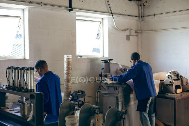 Rear view of two African American men working at a factory making cricket balls, one is feeding a piece of leather into a machine. — Stock Photo