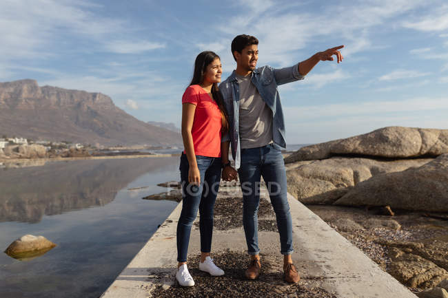 Vista frontale da vicino di una giovane coppia mista che si tiene per mano in piedi su un molo guardando verso il mare, l'uomo sta indicando la distanza — Foto stock