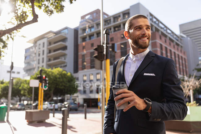 Вид спереди на улыбающегося молодого кавказца в куртке, стоящего на городской улице, держащего кофе на вынос и отводящего взгляд. Цифровая реклама на ходу . — стоковое фото
