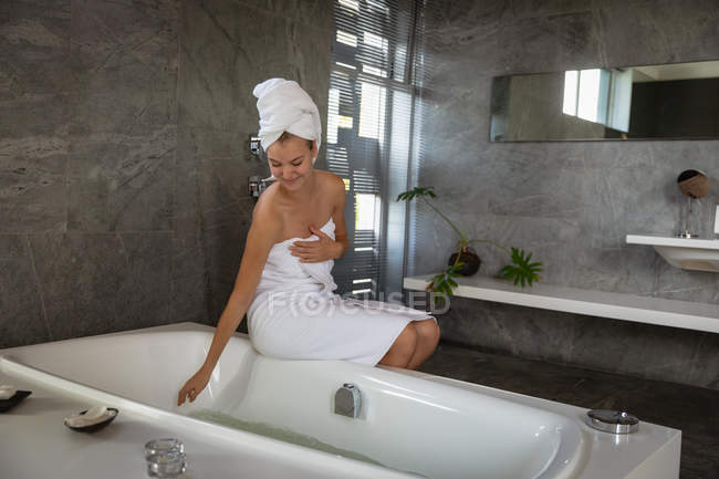 Vista frontal de uma jovem caucasiana vestindo uma toalha de banho e com o cabelo envolto em uma toalha, sentada na borda da banheira e tocando a água em um banheiro moderno . — Fotografia de Stock