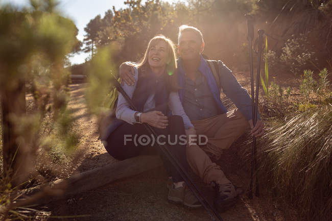 Front view of a mature Caucasian woman and man sitting on a fallen tree together holding Nordic walking sticks and admiring the scenery during a hike, backlit by sunlight — Stock Photo