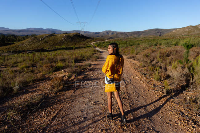 Vista laterale di una giovane donna di razza mista che cammina lungo un sentiero attraverso un paesaggio rurale soleggiato e si guarda intorno . — Foto stock