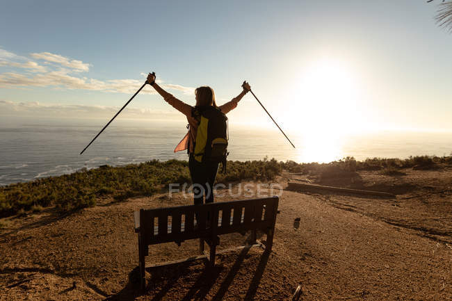 Rear view of a mature Caucasian woman wearing a backpack standing on a bench, raising her Nordic walking sticks in the air and enjoying the view of the coast at sunset during a hike. — Stock Photo