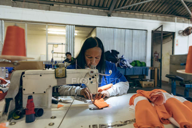 Vue de face d'une femme de race mixte d'âge moyen assise et travaillant à une machine à coudre dans une usine de vêtements de sport, avec un collègue travaillant à une machine à coudre en arrière-plan . — Photo de stock
