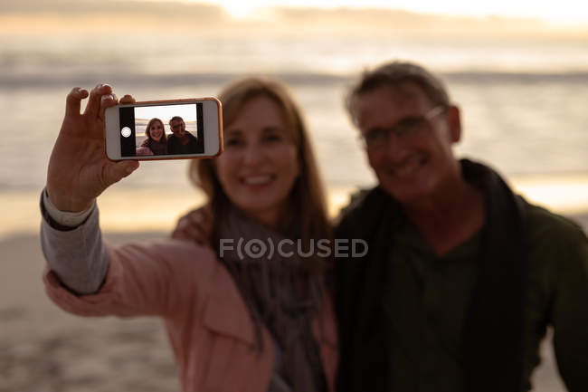 Вид спереди на взрослого белого мужчину и женщину, улыбающихся и делающих селфи со смартфоном на пляже перед морем на закате — стоковое фото